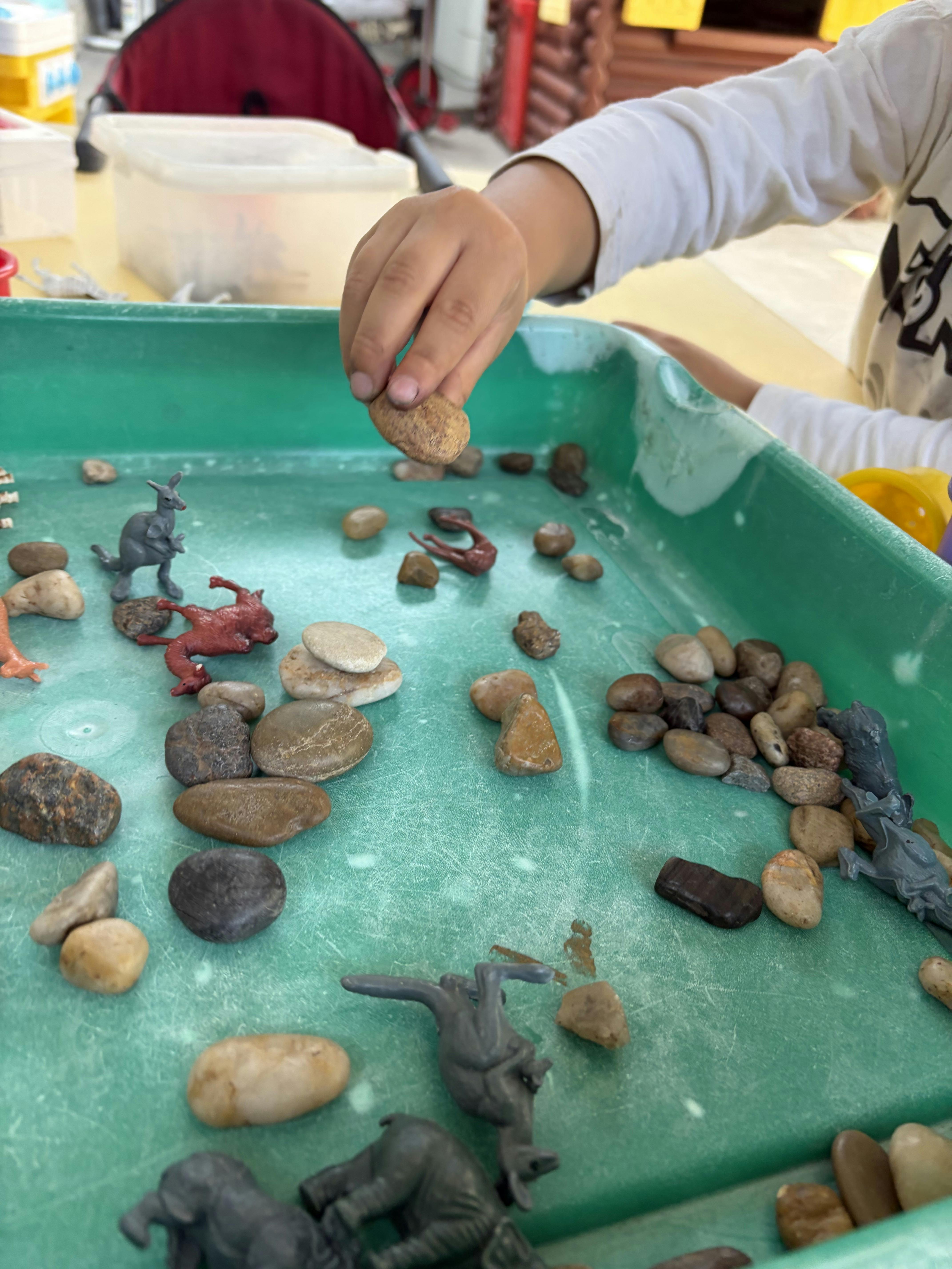 Child's hand holding a small rock over a green tray with toy animals