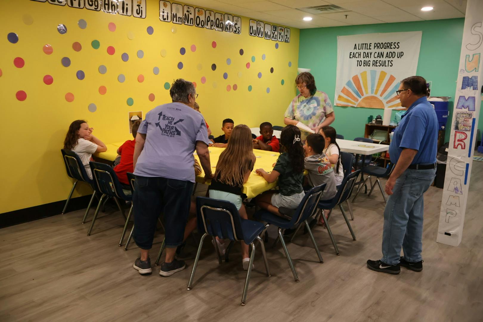 Adults assisting children at a table during a summer camp session