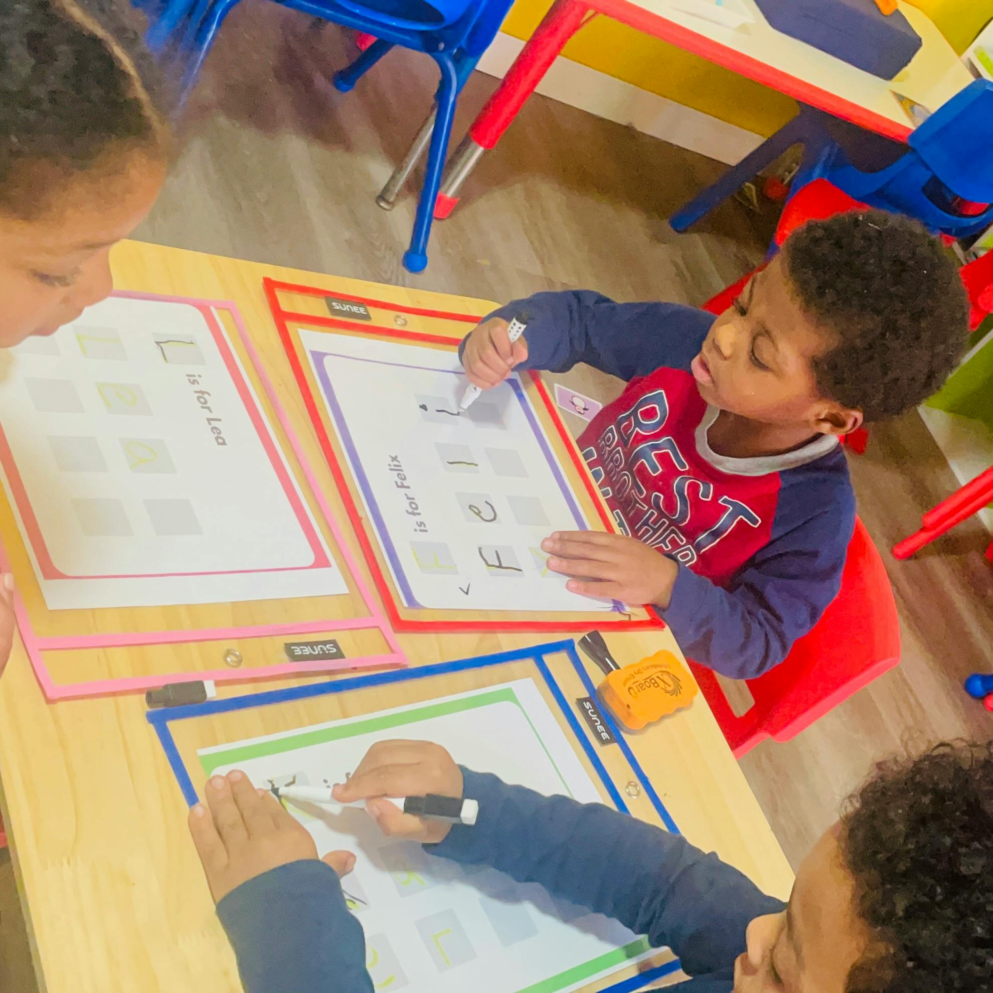 Three children in a classroom learning to write, tracing letters on worksheets