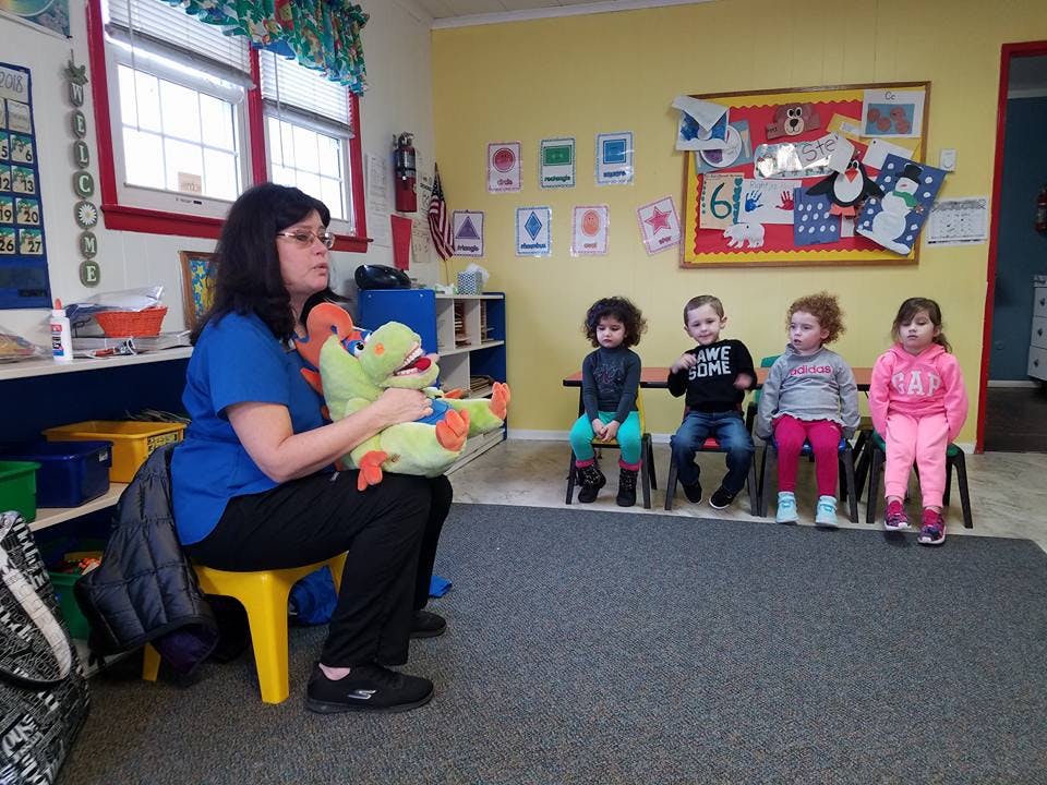 Person holding a stuffed animal in front of children sitting on chairs