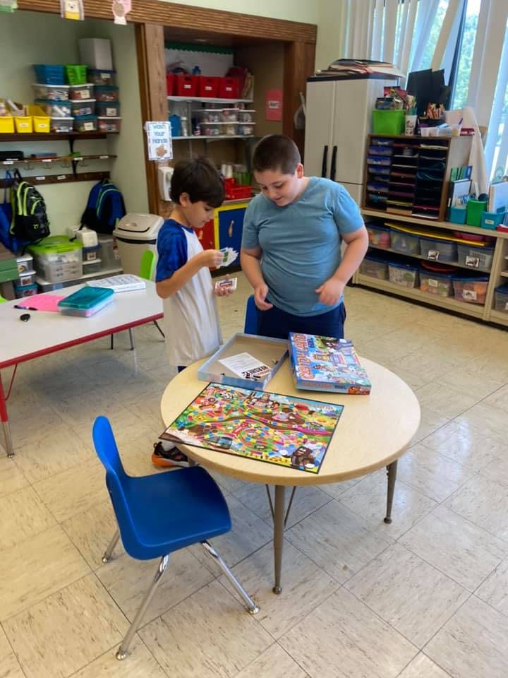 Two children standing at a table with a board game in a classroom
