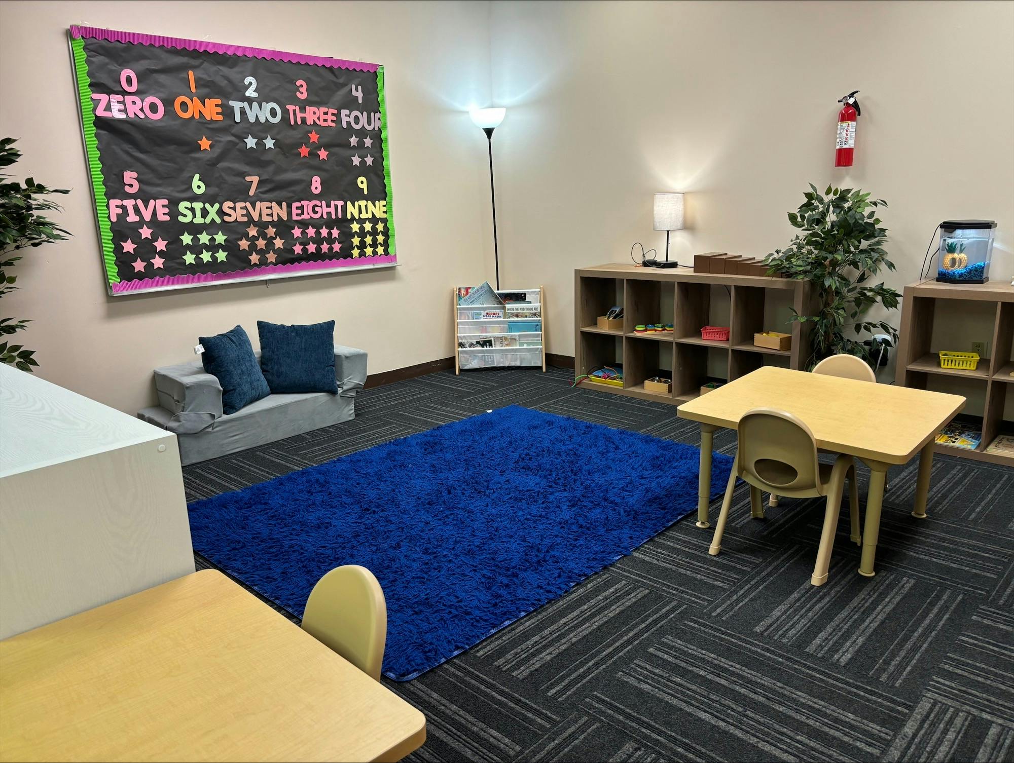 Classroom with a number chart, blue rug, small tables, and shelves