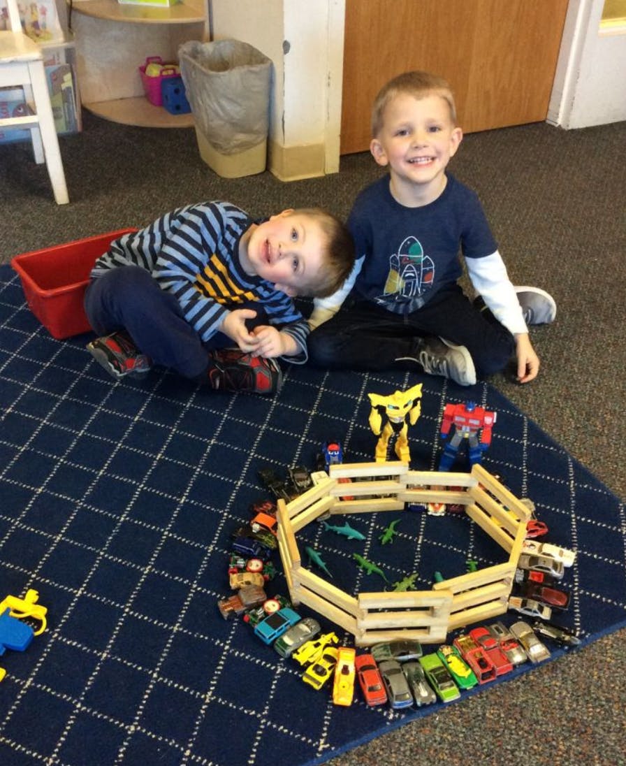 Two children on a rug with toy cars and a wooden fence