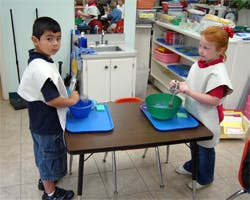 Two children in smocks standing at a table with bowls and trays