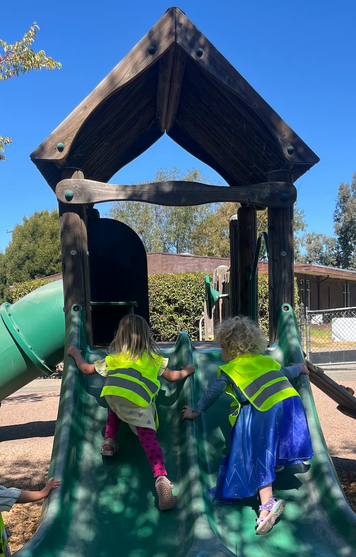 Two children wearing high-visibility vests climbing up a playground slide