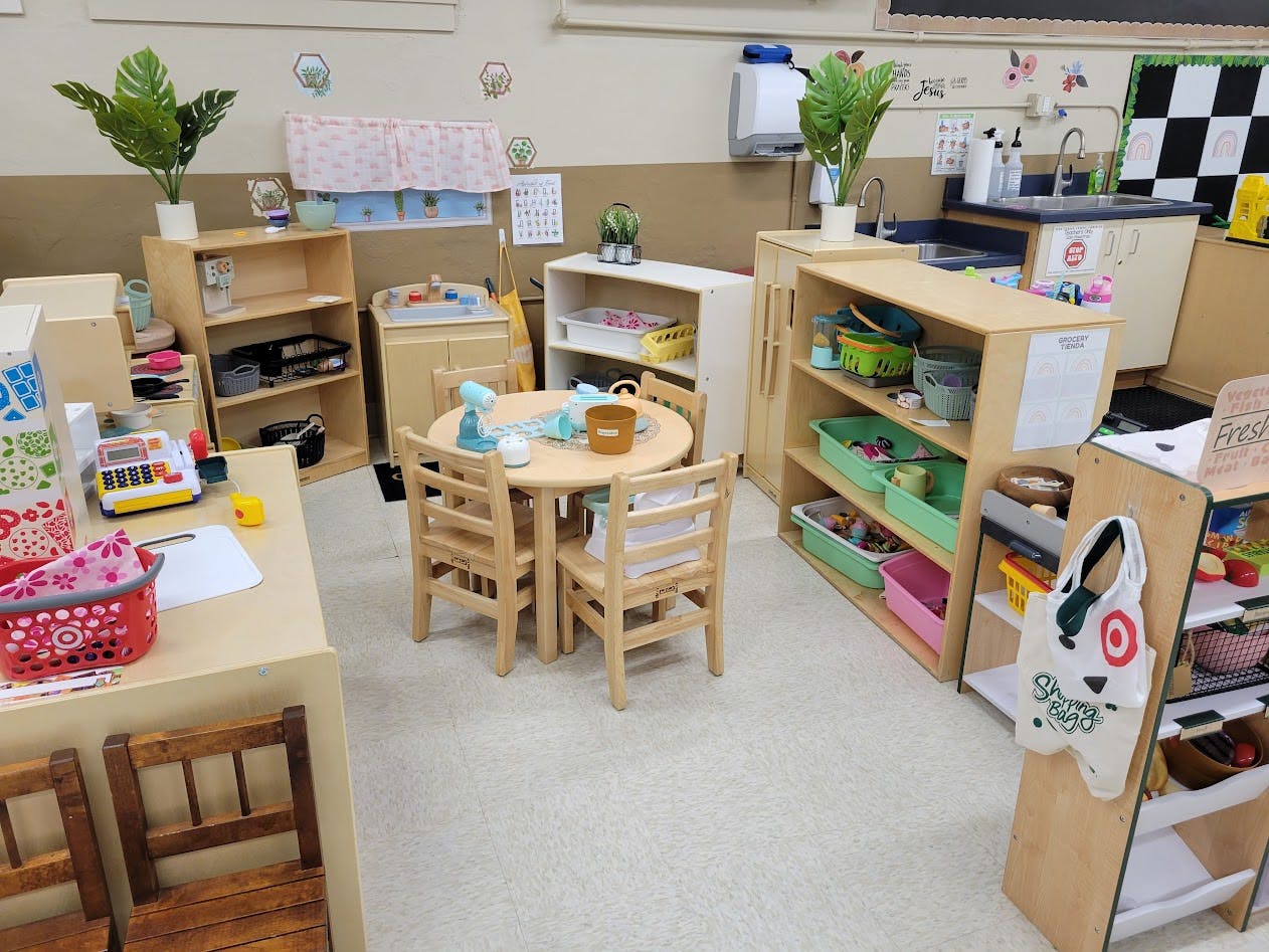 A classroom play area with a small round table and chairs, shelves, and pretend play items
