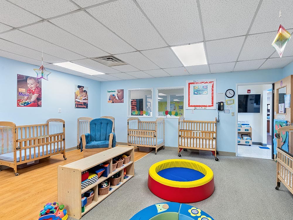A brightly lit nursery room with several wooden cribs and a blue rocking chair