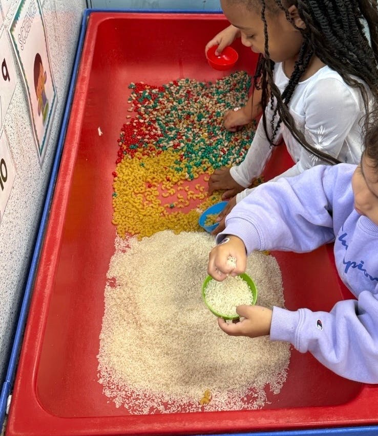 Two children playing with dried pasta and rice in a red bin