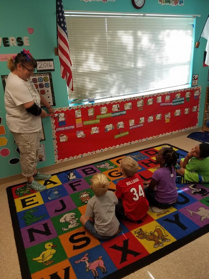 Adult standing in classroom with children sitting on alphabet rug