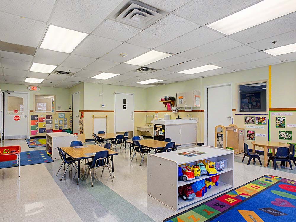 Empty classroom with small tables and chairs, toys on shelves, and an alphabet rug
