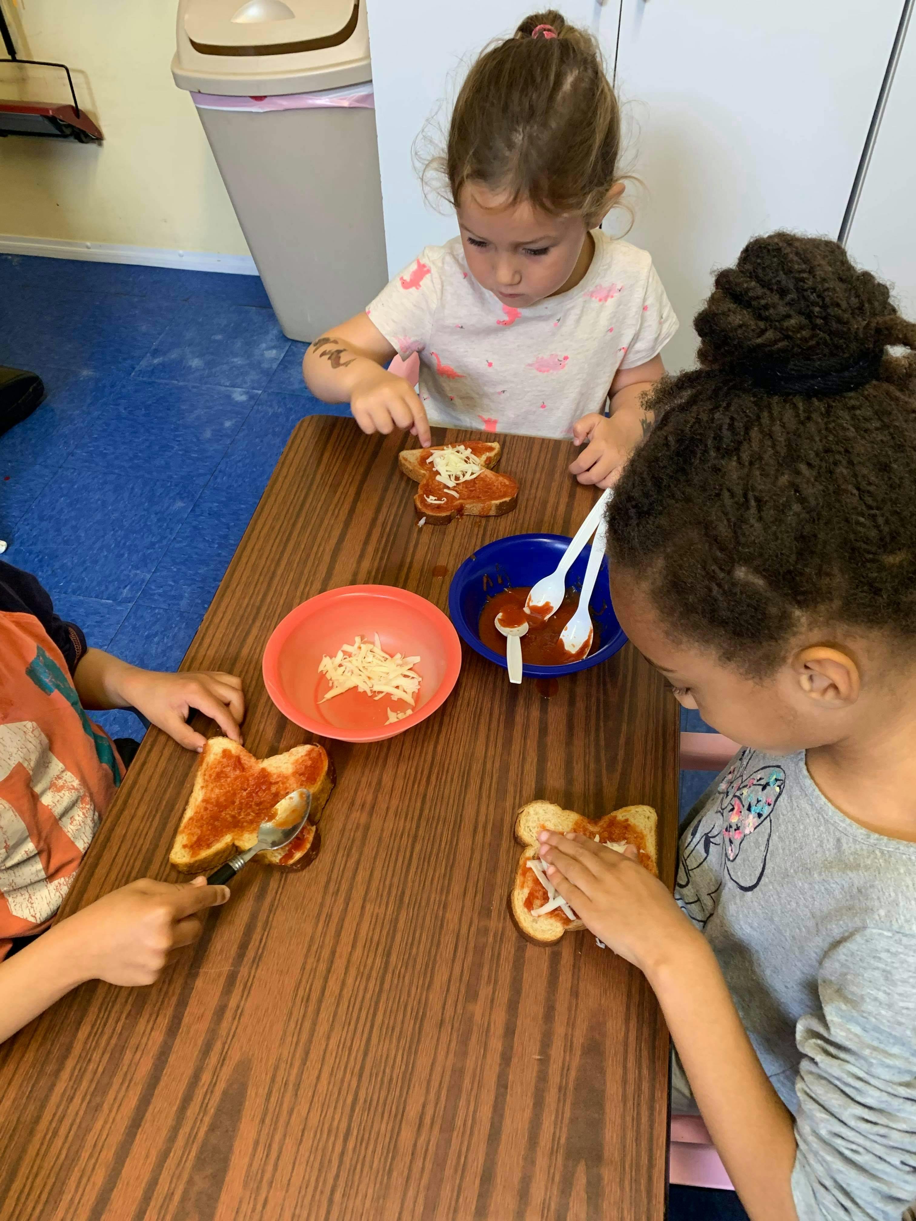 Three children making mini pizzas on toast at a table