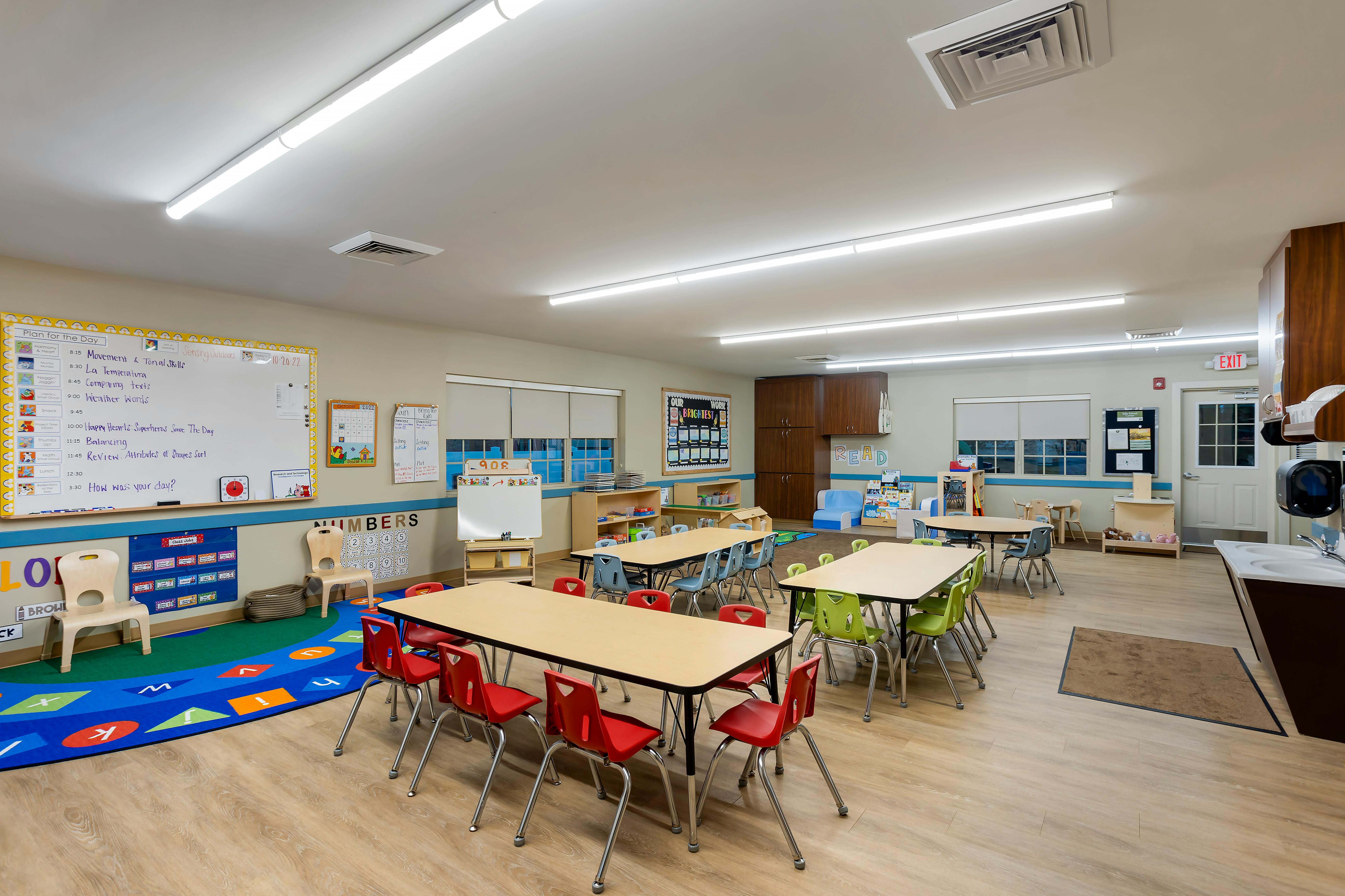 A brightly lit classroom with tables, chairs, a whiteboard, and a colorful rug