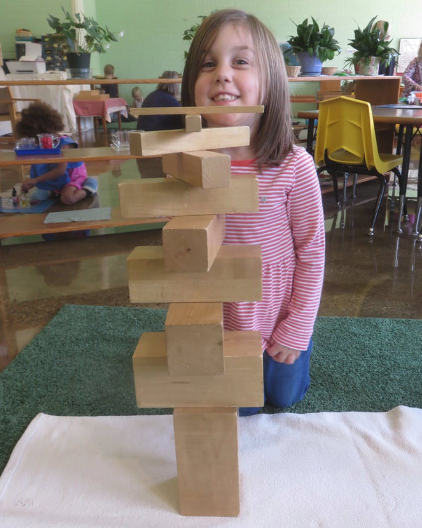 Child kneeling behind a tall tower of wooden blocks in a classroom