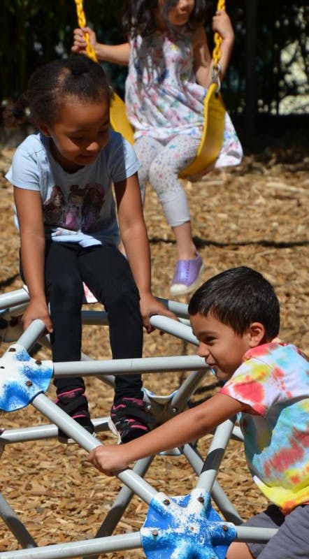 Three children playing on a playground, one on a swing, two on a climbing dome