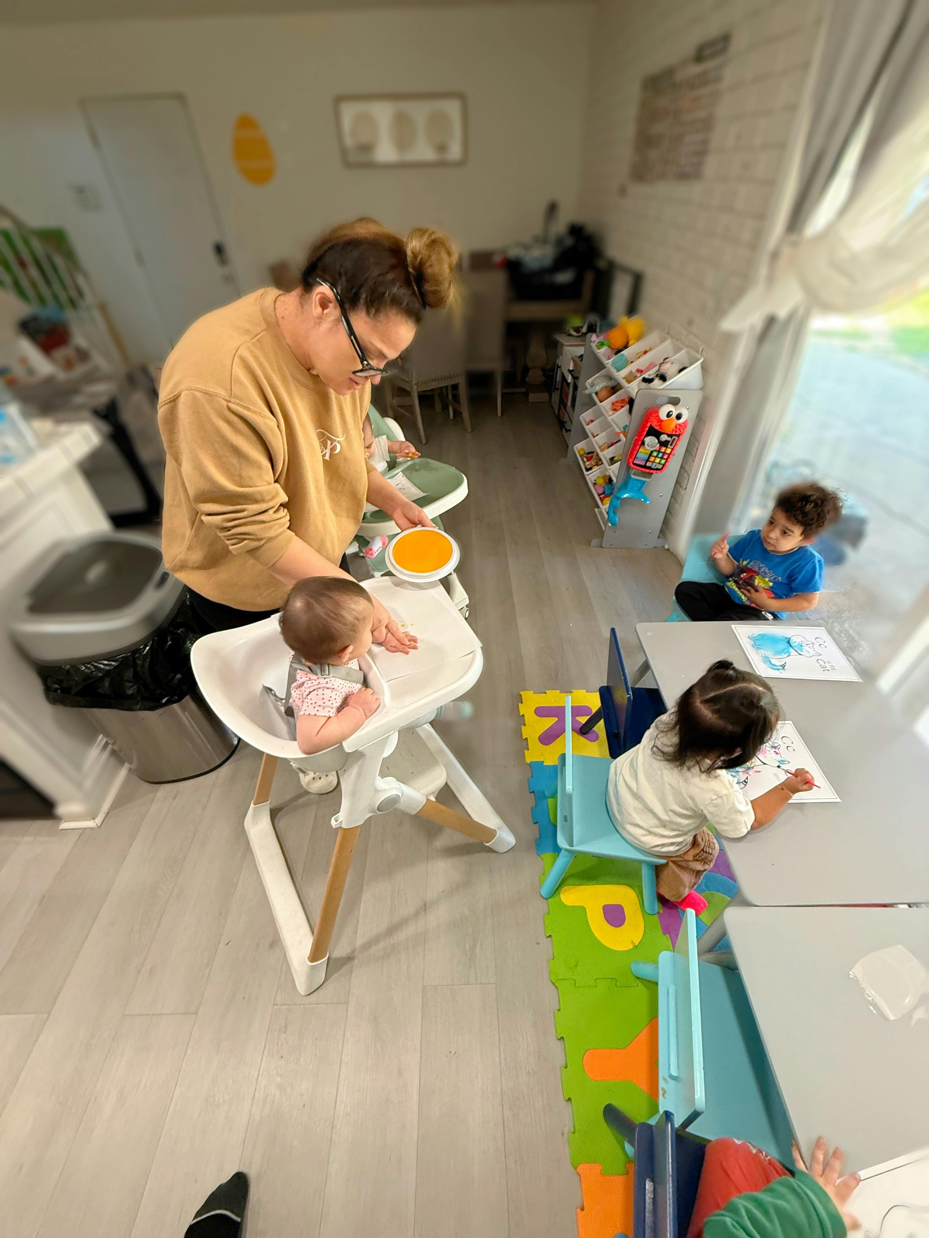 Person feeding a baby in a high chair while two children draw at small desks