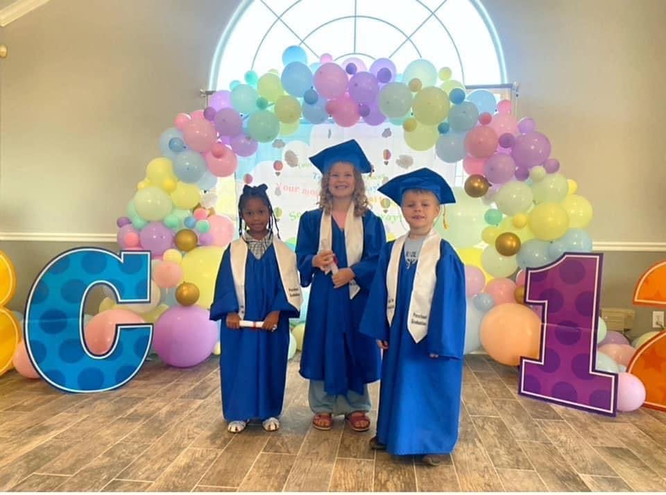 Three children in blue graduation gowns and caps standing in front of balloons