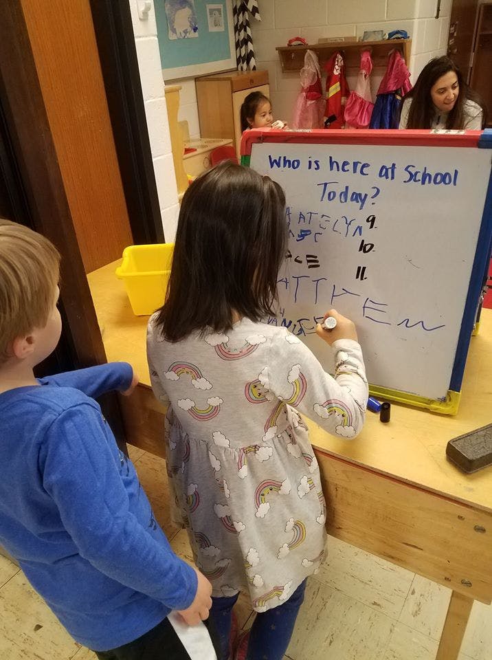 Child writing on a whiteboard that says "Who is here at School Today?"