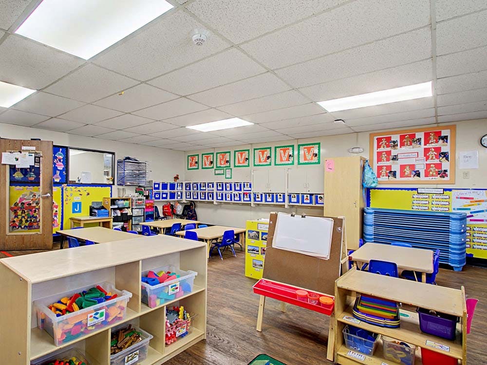 Empty classroom with small tables and chairs, shelves with toys, and an easel