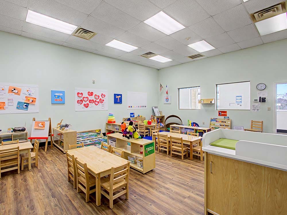 Classroom with small wooden tables and chairs, shelves of toys, and art on the walls