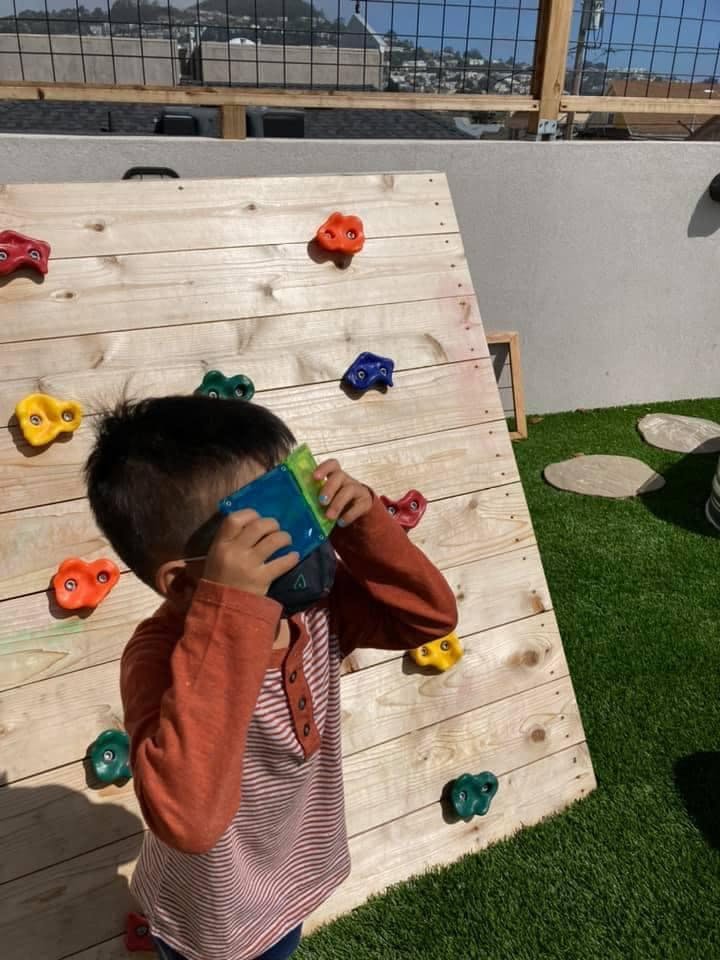Child holding a blue object to their face next to a climbing wall