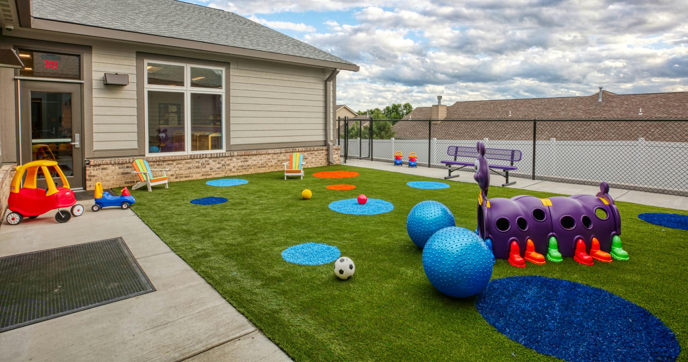 An outdoor play area with artificial turf, various colorful toys, and a building