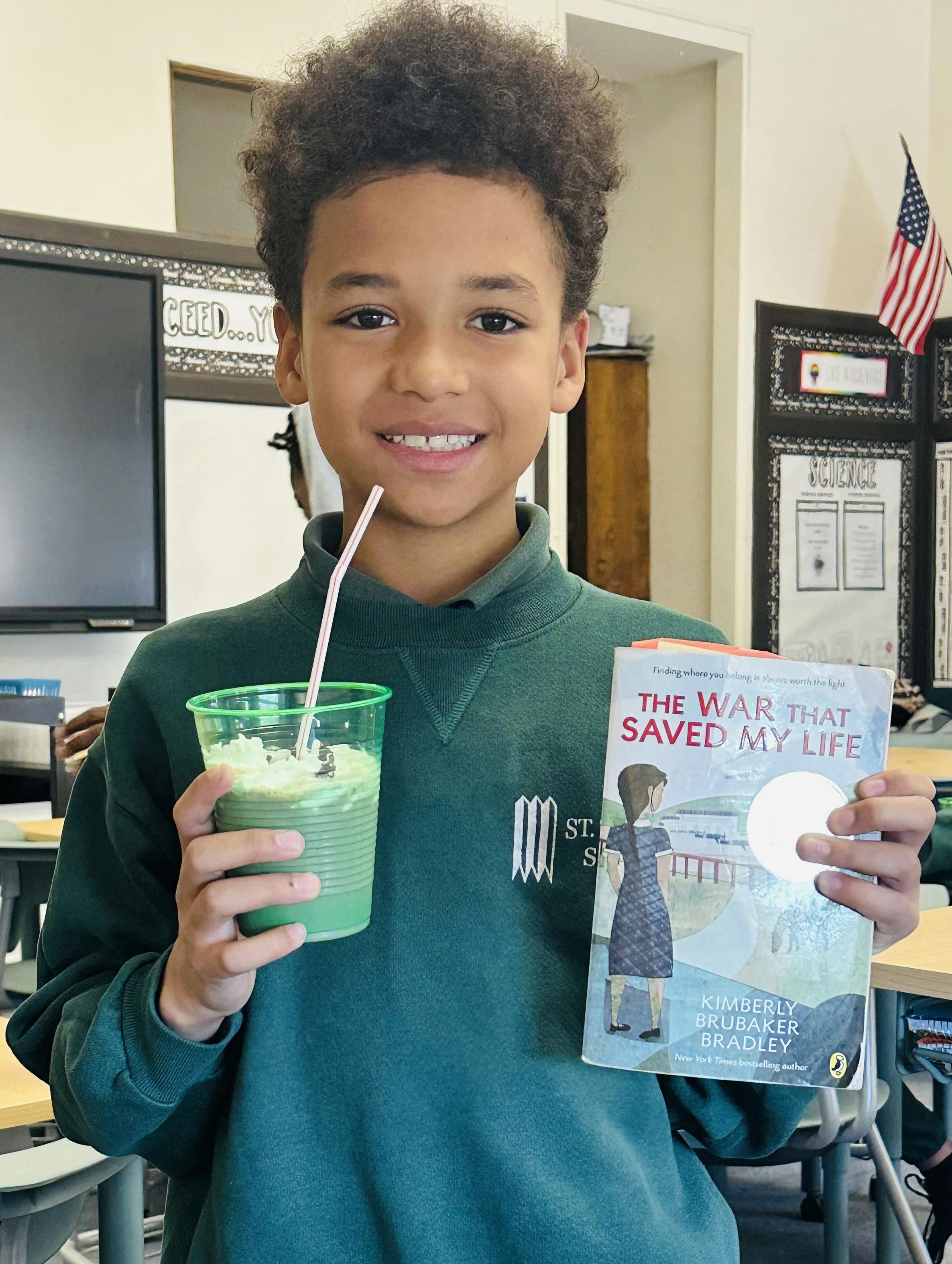 Child holding a green drink and a book titled "The War That Saved My Life"