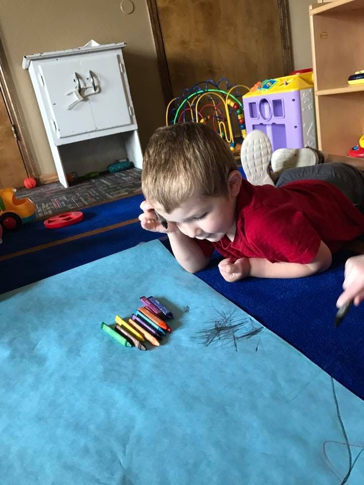 Child lying on floor drawing on blue paper with crayons