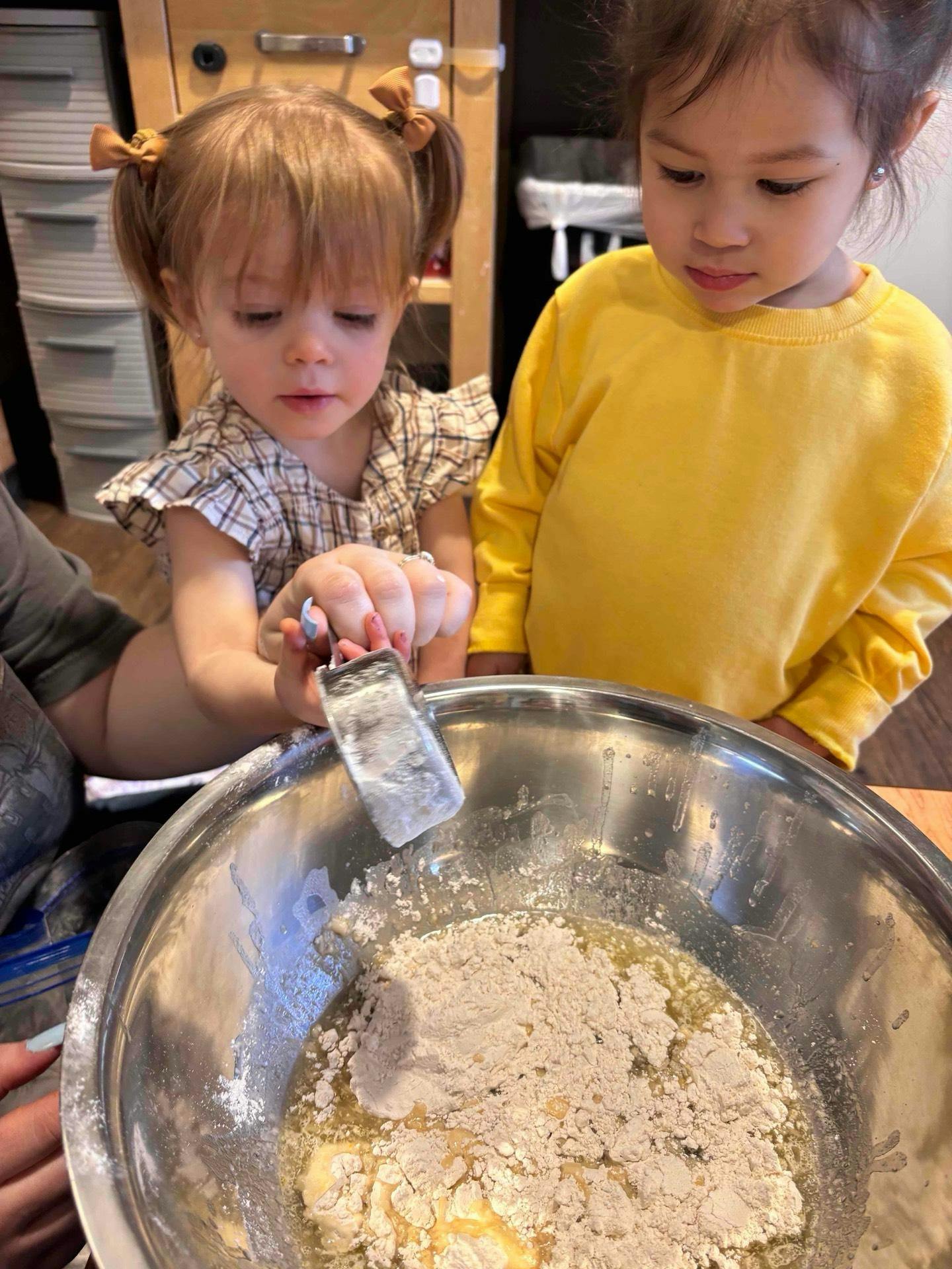 Two children pouring ingredients into a metal mixing bowl