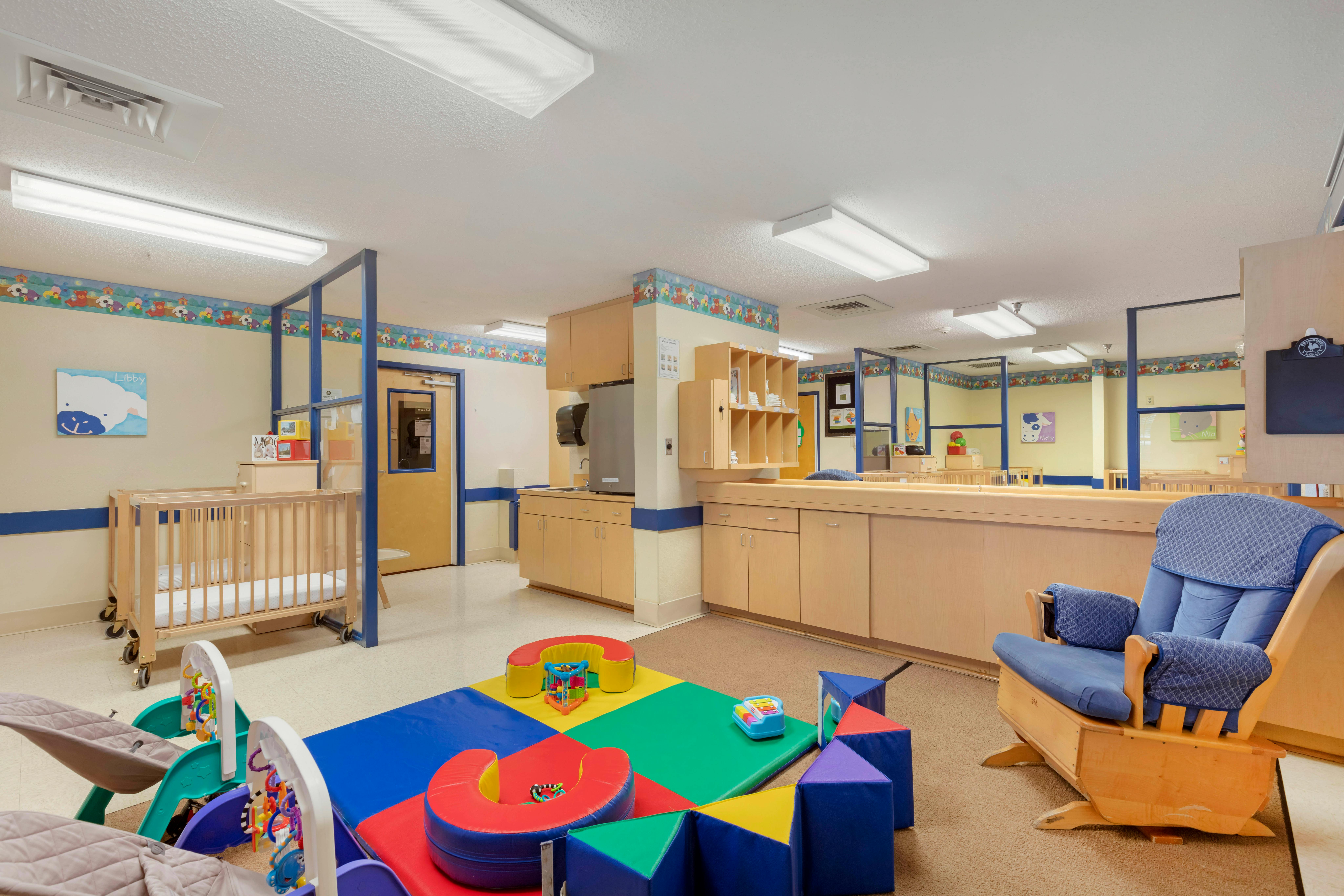 A colorful childcare room with cribs, playmats, and a rocking chair