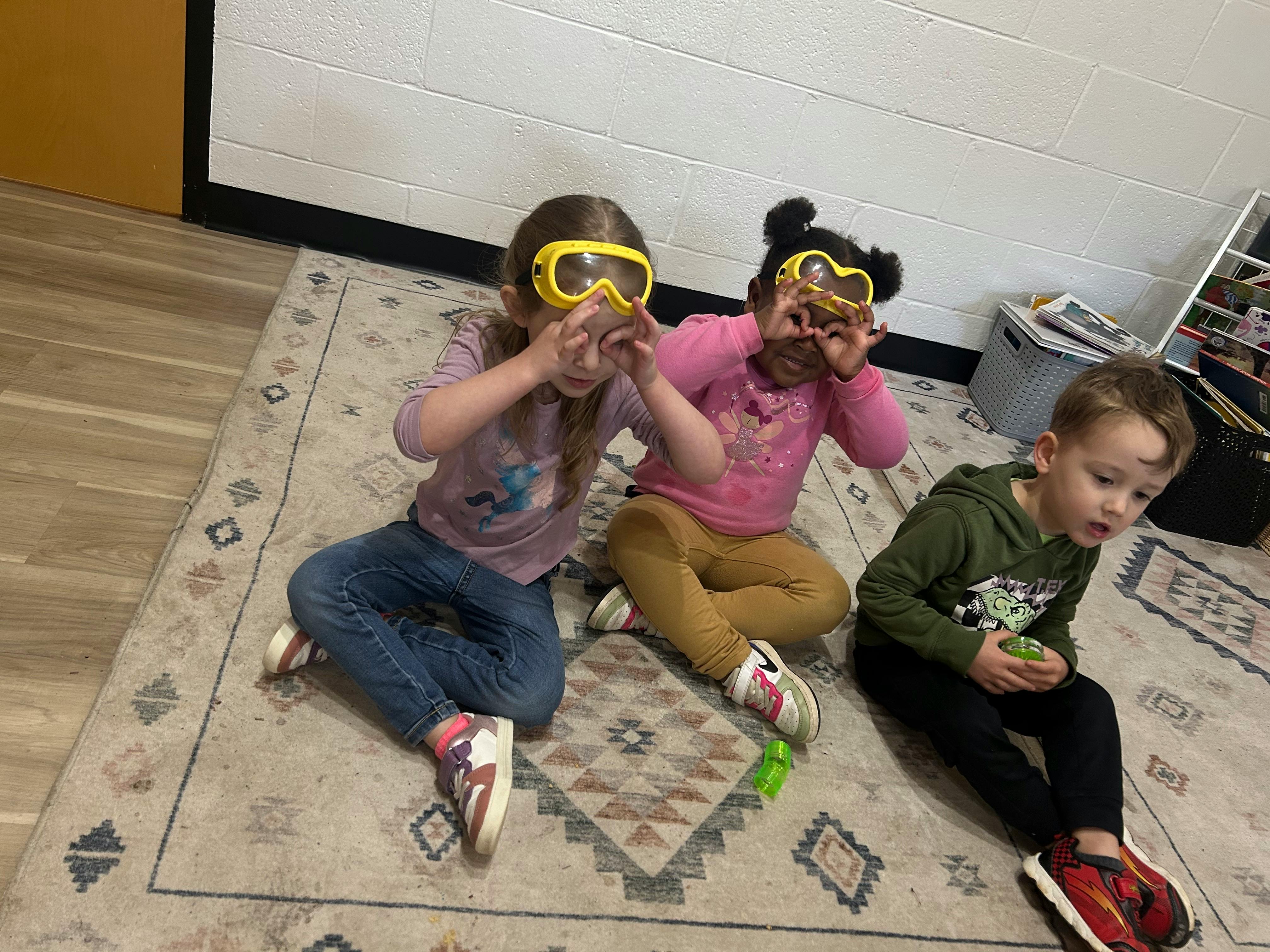 Three children lying on a rug, two wearing yellow safety goggles