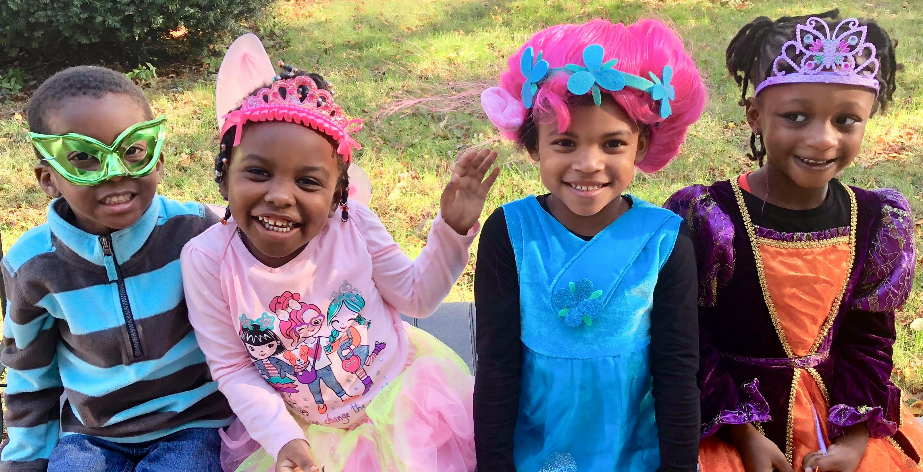 Four children in costumes, one wearing a green mask, one a pink tiara, one a pink wig