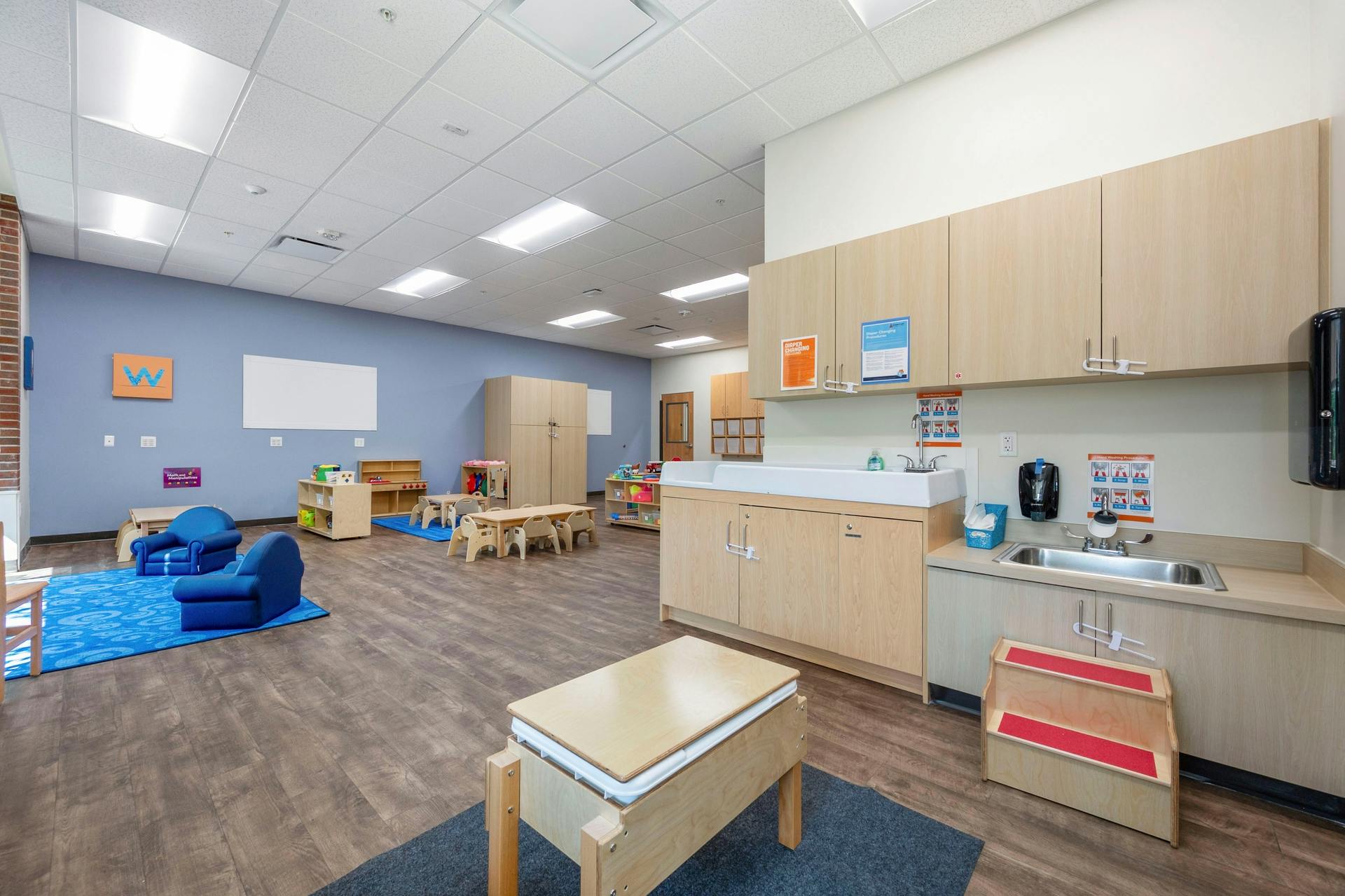 Classroom with wooden floors, blue walls, a changing station, sink, and small furniture