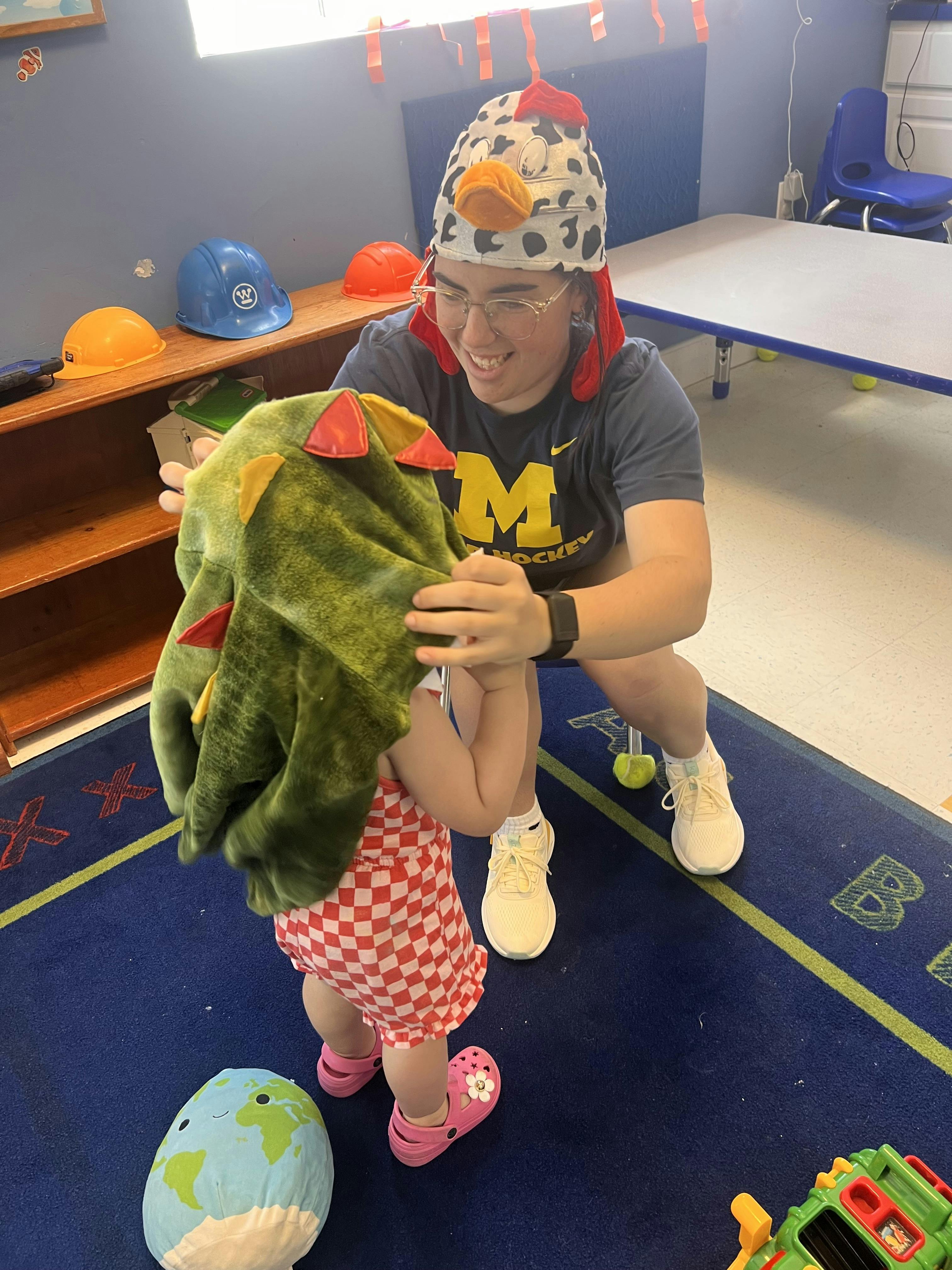 Person wearing a chicken hat putting a green dinosaur hood on a child
