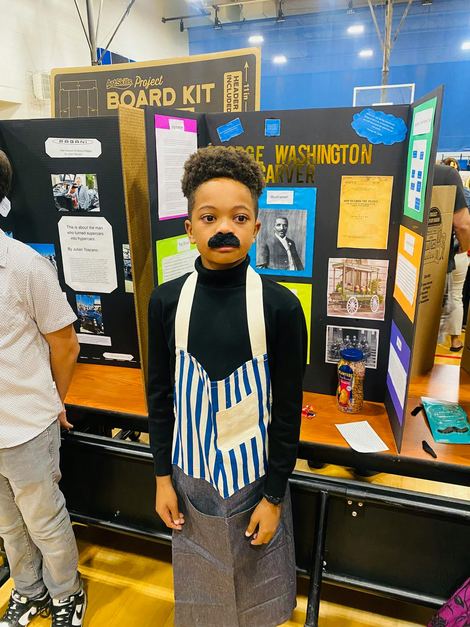 Child with a fake mustache and apron standing in front of a project board