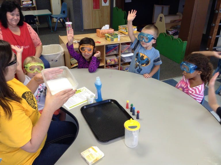 Adult and children in safety goggles at a table with science experiment supplies