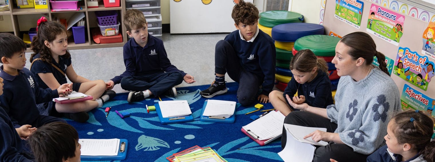 Teacher and children sitting on a rug in a classroom, looking at papers