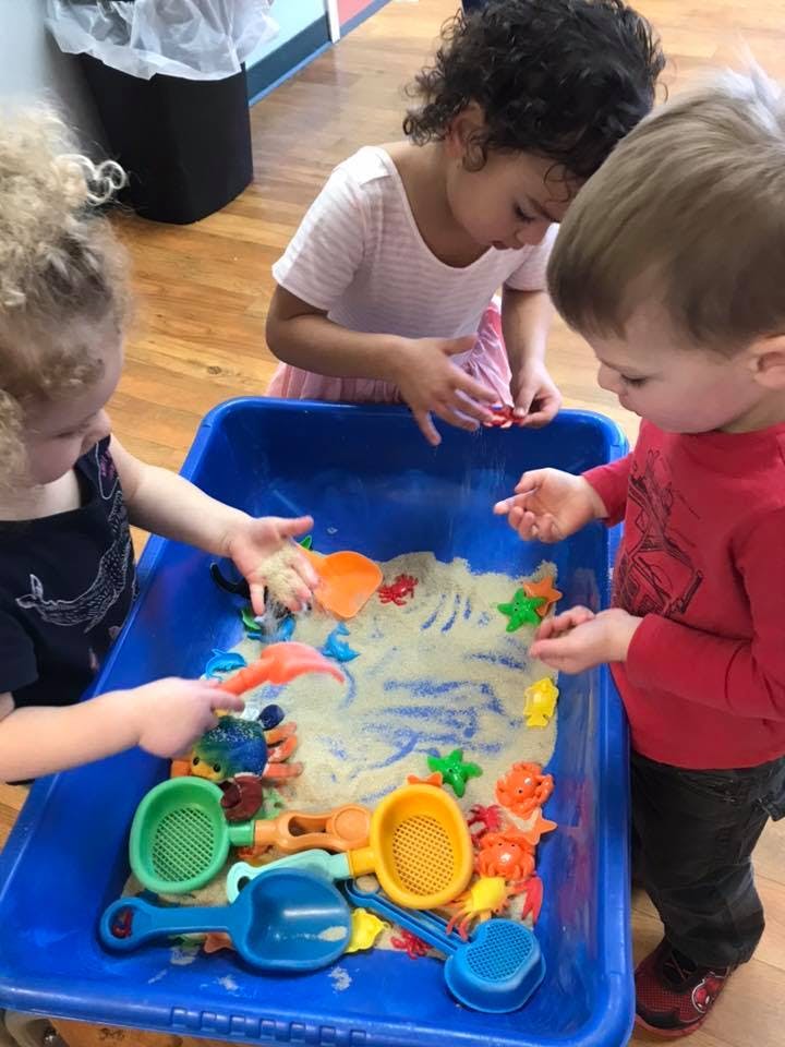 Three children playing with sand and toys in a blue bin
