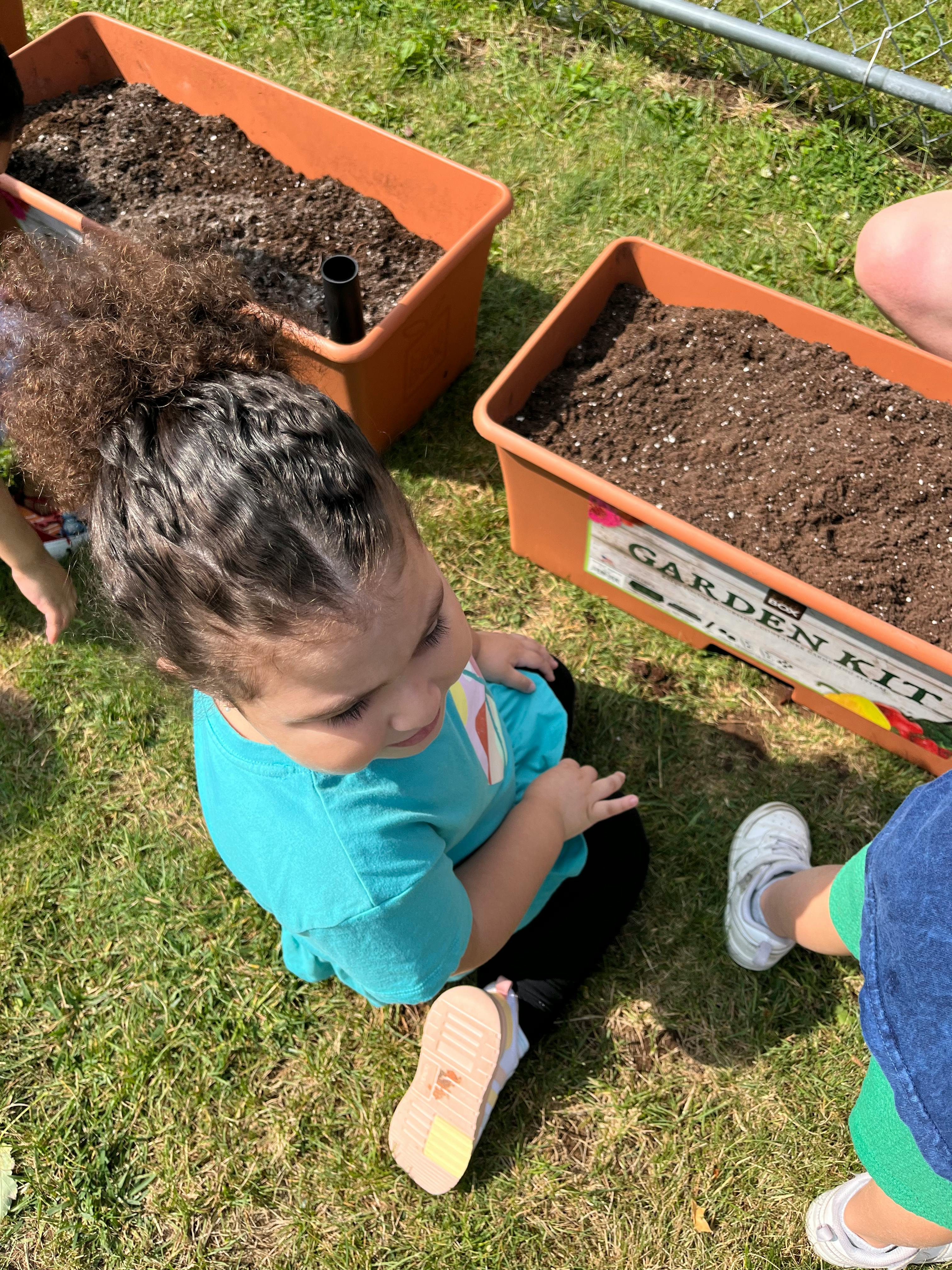Child sitting on grass next to garden planters filled with soil