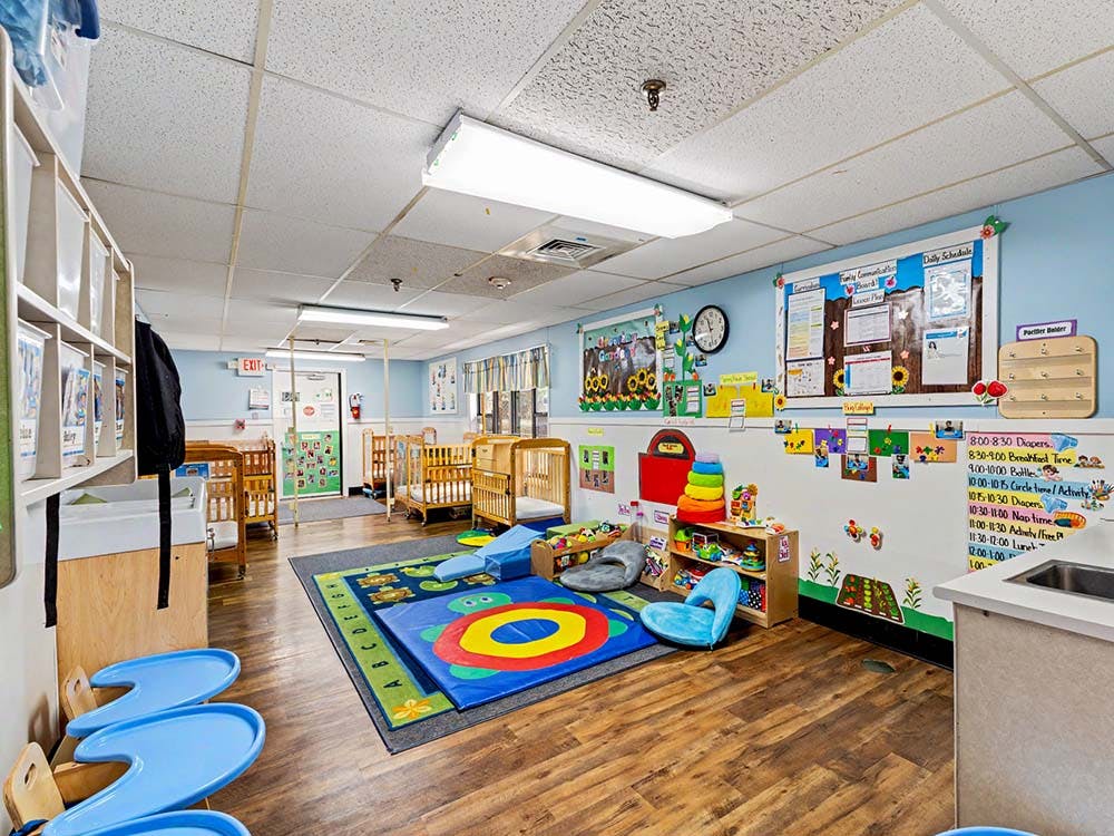 Classroom with cribs, play rug, and activity schedule on the wall