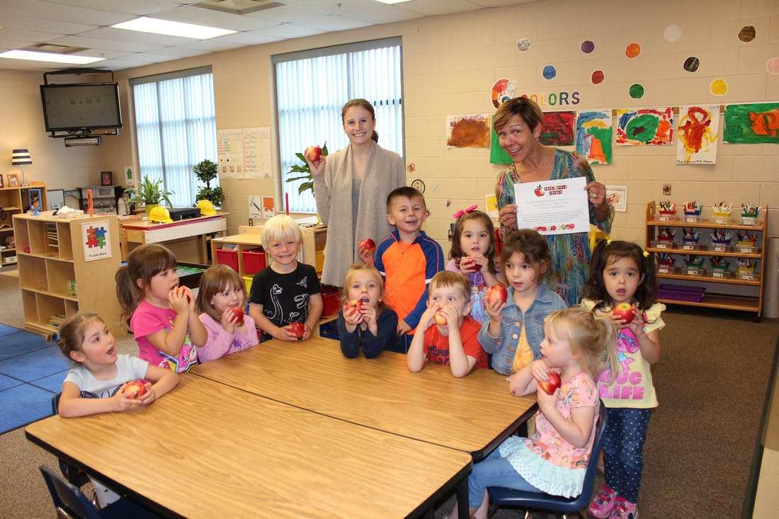 Two adults and multiple children holding apples in a classroom