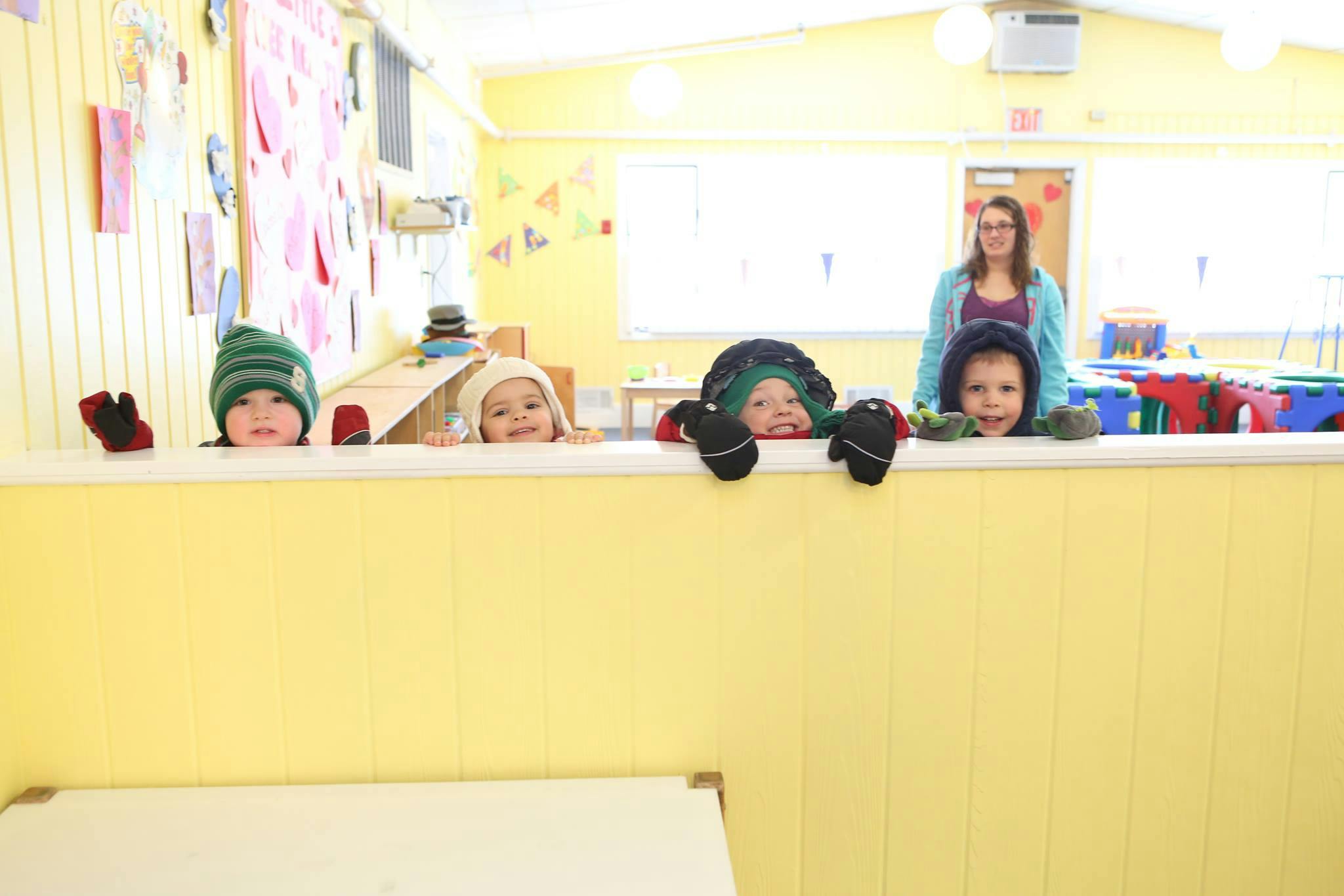 Four children in hats peeking over a yellow wall in a classroom, with a person standing behind them