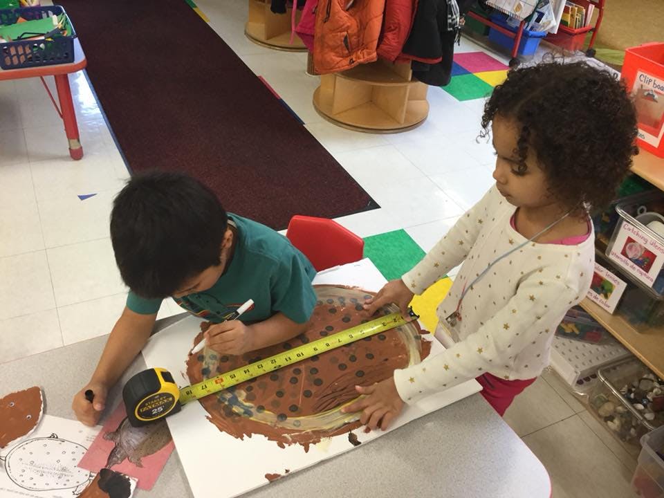 Two children measuring a brown painted shape with a tape measure