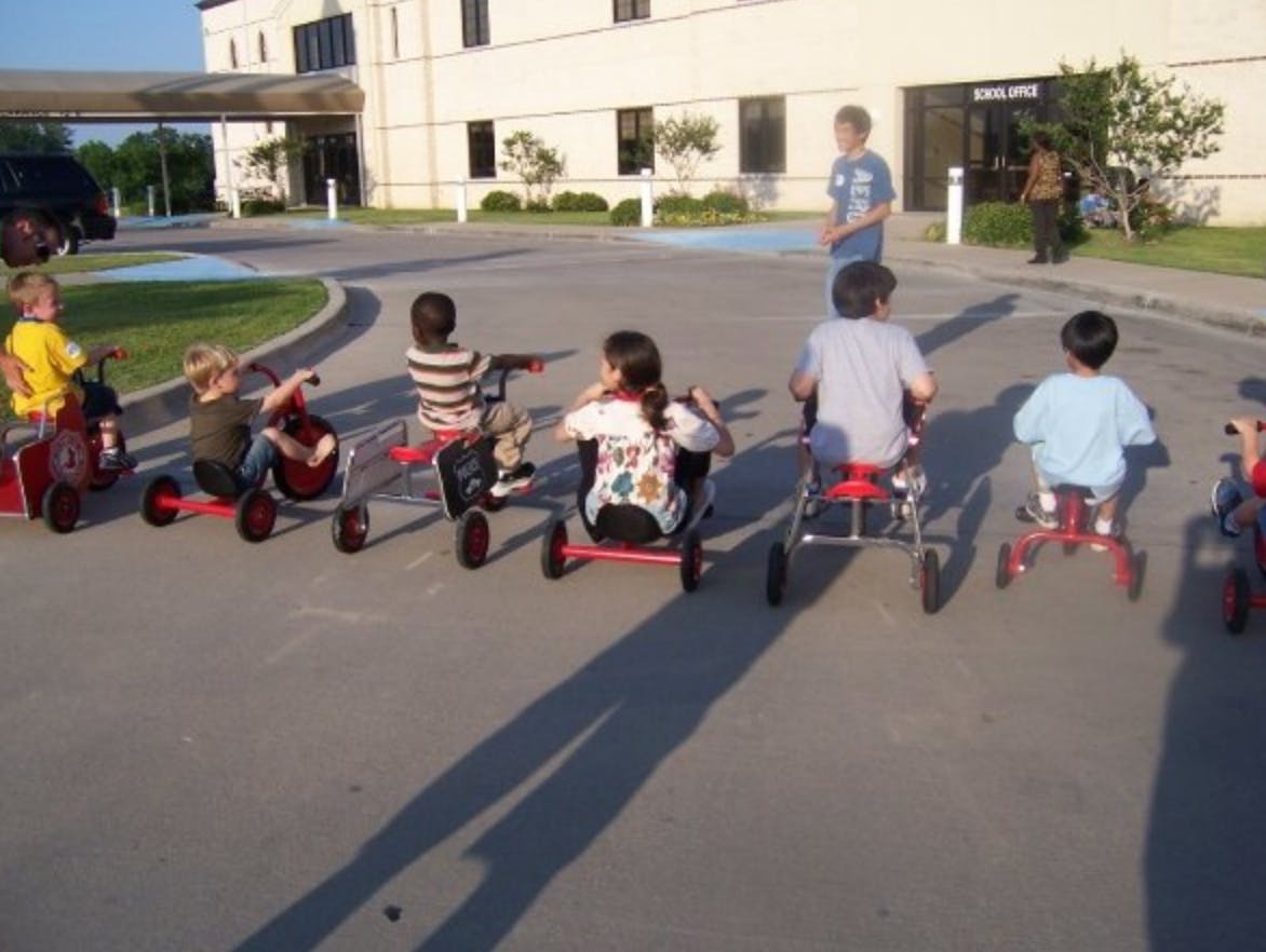 Children riding tricycles outdoors with an adult standing nearby