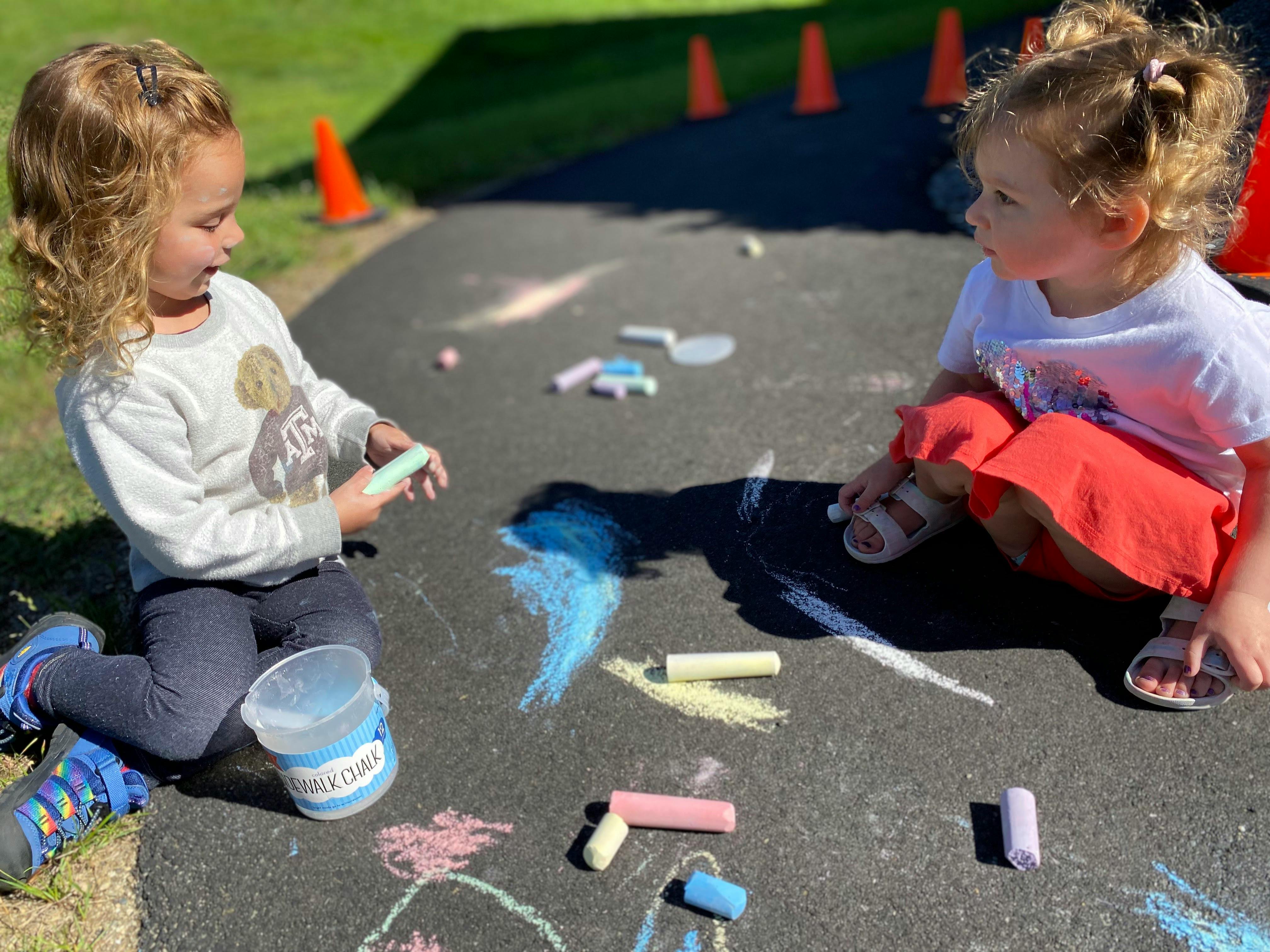 Two children sitting on pavement drawing with sidewalk chalk