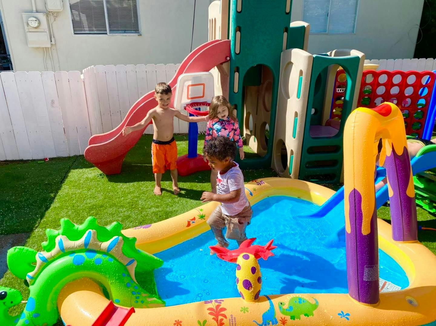 Three children playing in an outdoor area with a water slide and inflatable pool