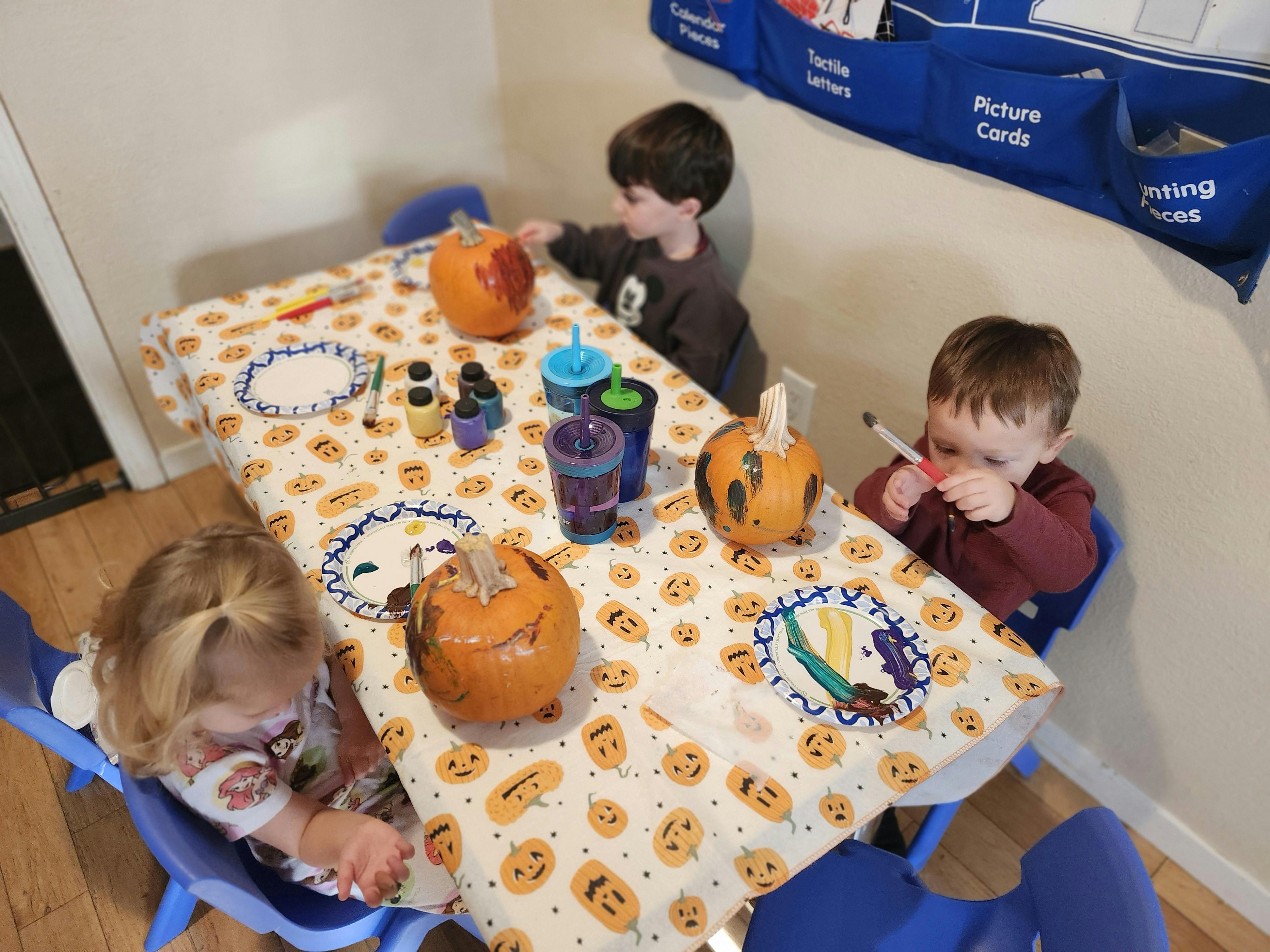 Three children sitting at a table with pumpkins, paint, and brushes