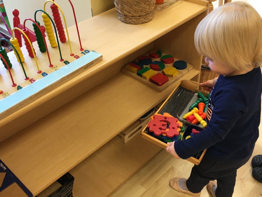 Child holding a tray of colorful toys next to a shelf with other toys