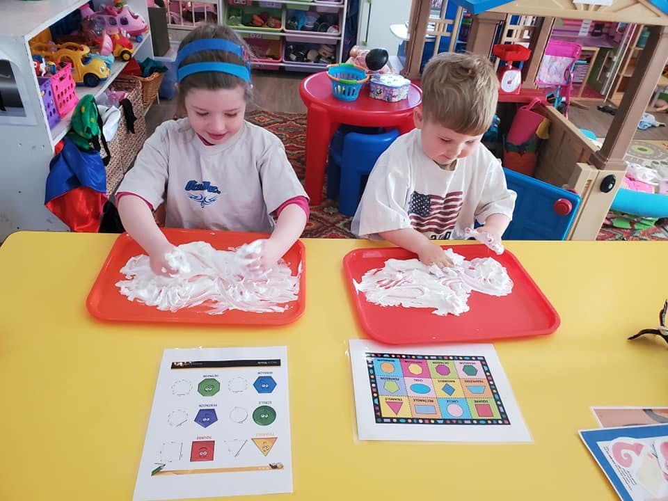 Two children playing with shaving cream on red trays at a yellow table