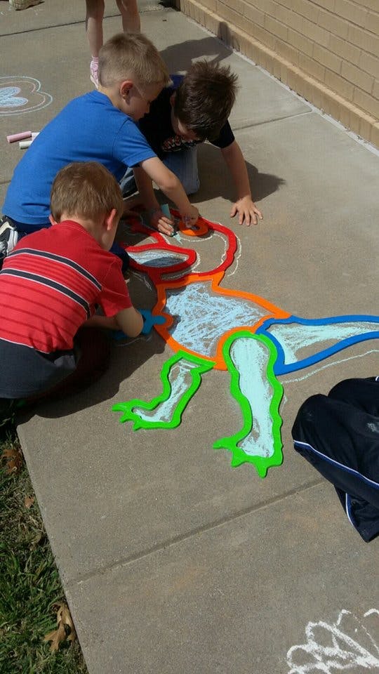 Three children drawing a dinosaur with chalk on a sidewalk