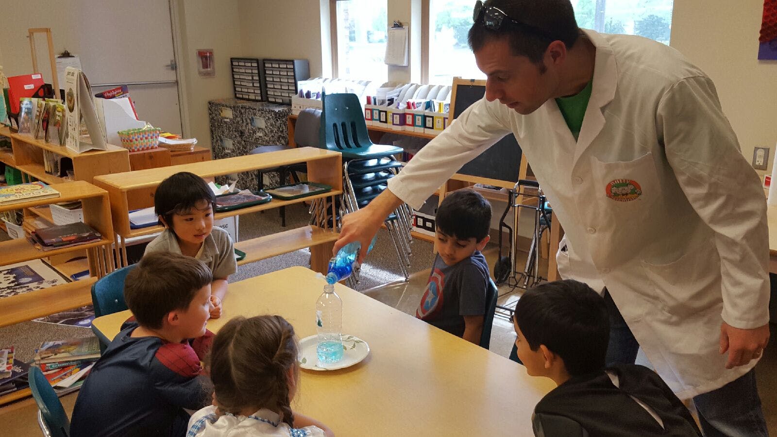 Person in lab coat pouring liquid for children at a table in a classroom